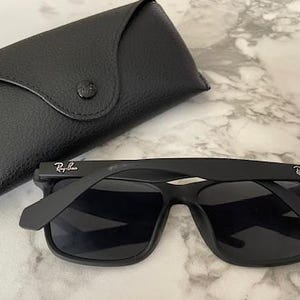 Pu&ograve; includere: Occhiali da sole neri con custodia nera abbinata. Gli occhiali hanno una montatura quadrata e il logo Ray-Ban sull'asta. La custodia &egrave; in pelle e ha una chiusura a bottone. Il logo Ray-Ban &egrave; visibile anche sulla custodia.
