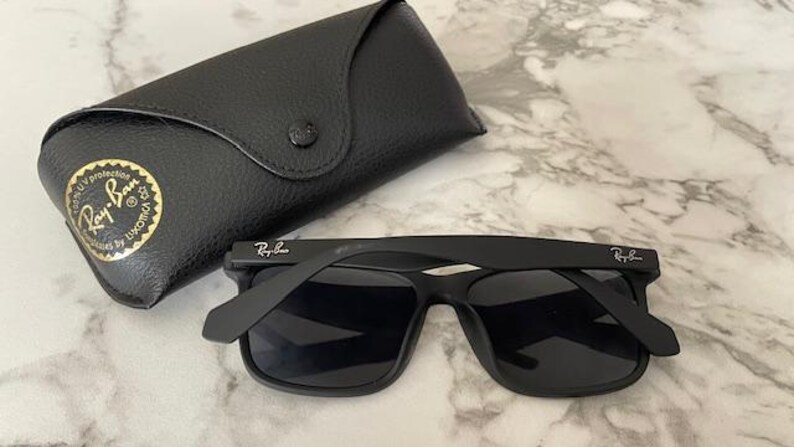 Pu&ograve; includere: Occhiali da sole neri con custodia nera abbinata. Gli occhiali hanno una montatura quadrata e il logo Ray-Ban sull'asta. La custodia &egrave; in pelle e ha una chiusura a bottone. Il logo Ray-Ban &egrave; visibile anche sulla custodia.