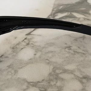May include: Close-up of black sunglasses with a sleek, modern design. The sunglasses feature a curved arm and a dark lens, suggesting a sporty or casual style. The frame appears to be made of a durable material.