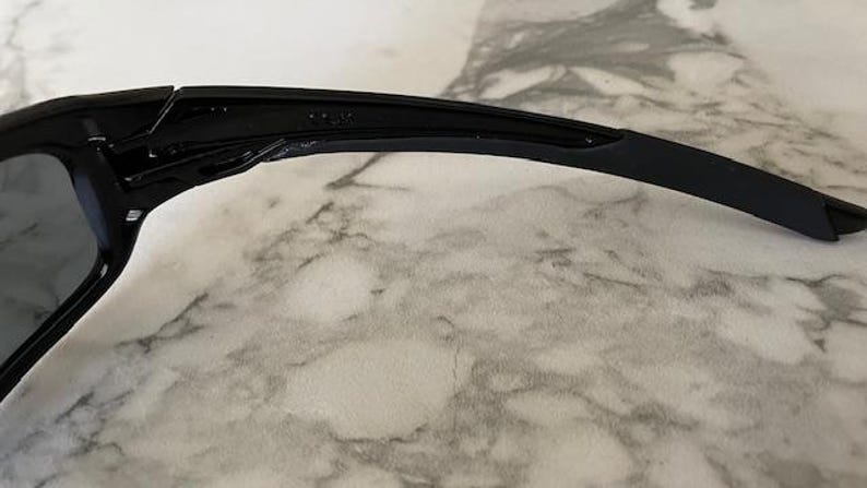 May include: Close-up of black sunglasses with a sleek, modern design. The sunglasses feature a curved arm and a dark lens, suggesting a sporty or casual style. The frame appears to be made of a durable material.