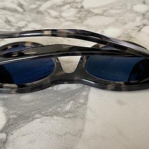 Puede incluir: Gafas de sol con montura con estampado de carey y lentes azul oscuro. Las gafas de sol est&aacute;n plegadas, revelando el marco interior y las bisagras. La montura tiene un patr&oacute;n moteado de negro, gris y beige.