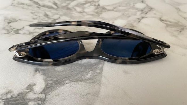 Puede incluir: Gafas de sol con montura con estampado de carey y lentes azul oscuro. Las gafas de sol est&aacute;n plegadas, revelando el marco interior y las bisagras. La montura tiene un patr&oacute;n moteado de negro, gris y beige.