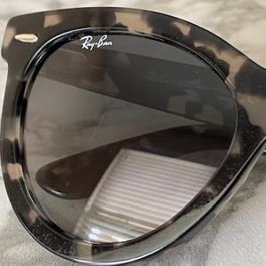 Puede incluir: Primer plano de gafas de sol Ray-Ban con montura con estampado de carey. Las lentes son oscuras y el logotipo de Ray-Ban es visible en la esquina superior izquierda de la montura. Las gafas de sol tienen una forma cl&aacute;sica y redondeada.