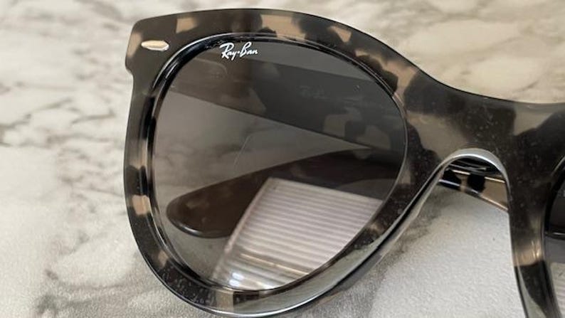 Puede incluir: Primer plano de gafas de sol Ray-Ban con montura con estampado de carey. Las lentes son oscuras y el logotipo de Ray-Ban es visible en la esquina superior izquierda de la montura. Las gafas de sol tienen una forma cl&aacute;sica y redondeada.