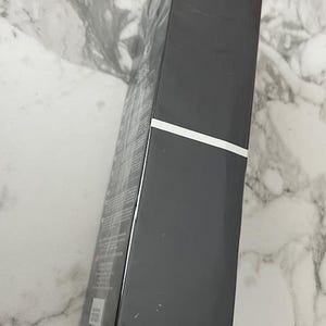 May include: A black rectangular box with a white stripe, wrapped in clear plastic. The box is angled, revealing text and graphics on the side. The background is a white and gray marble pattern.
