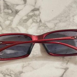 May include: Red-framed sunglasses with dark lenses. The sunglasses have a rectangular shape and a glossy finish. The arms of the sunglasses are thin and curve around the ears. The Oakley logo is visible on the temples.