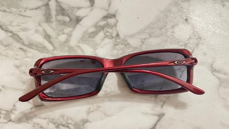 May include: Red-framed sunglasses with dark lenses. The sunglasses have a rectangular shape and a glossy finish. The arms of the sunglasses are thin and curve around the ears. The Oakley logo is visible on the temples.