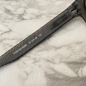 May include: Close-up of Holbrook sunglasses with a dark gray frame. The arm of the sunglasses displays the text "HOLBROOK" and the numbers "OO9102-X555 57 18 137". The lenses are a dark color.