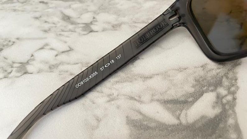 May include: Close-up of Holbrook sunglasses with a dark gray frame. The arm of the sunglasses displays the text "HOLBROOK" and the numbers "OO9102-X555 57 18 137". The lenses are a dark color.