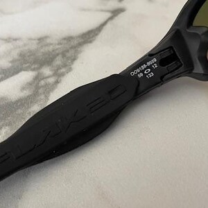 Puede incluir: Primer plano de gafas de sol negras con el logotipo FLAKED en la patilla. Las lentes tienen un tinte degradado. En la patilla se lee: OO9188-8659 59 12 133.