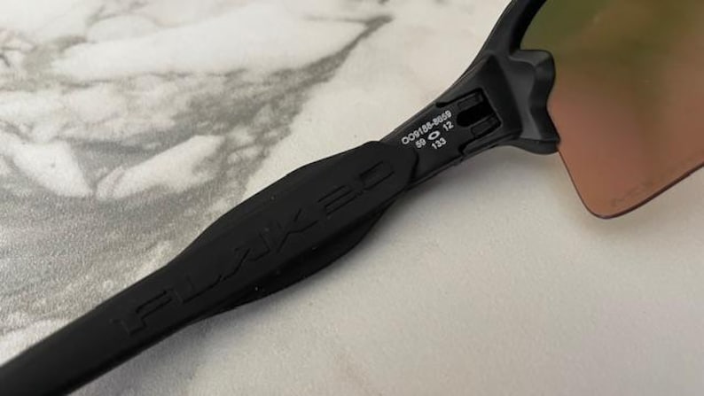 Puede incluir: Primer plano de gafas de sol negras con el logotipo FLAKED en la patilla. Las lentes tienen un tinte degradado. En la patilla se lee: OO9188-8659 59 12 133.