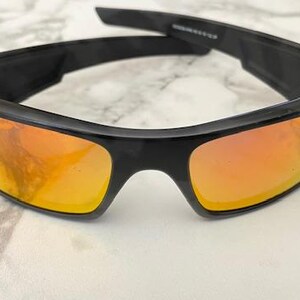 Puede incluir: Gafas de sol negras con lentes de color naranja. Las gafas tienen un dise&ntilde;o deportivo con una montura gruesa y rectangular. Las lentes reflejan un tono c&aacute;lido y dorado. Las patillas de las gafas tambi&eacute;n son negras.