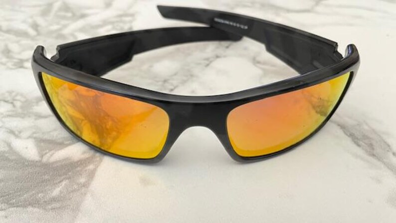 Puede incluir: Gafas de sol negras con lentes de color naranja. Las gafas tienen un dise&ntilde;o deportivo con una montura gruesa y rectangular. Las lentes reflejan un tono c&aacute;lido y dorado. Las patillas de las gafas tambi&eacute;n son negras.