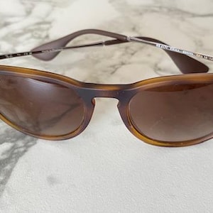 May include: Brown sunglasses with a tortoiseshell frame and gradient lenses. The sunglasses have a classic design with a subtle cat-eye shape. The Ray-Ban logo is visible on the frame.