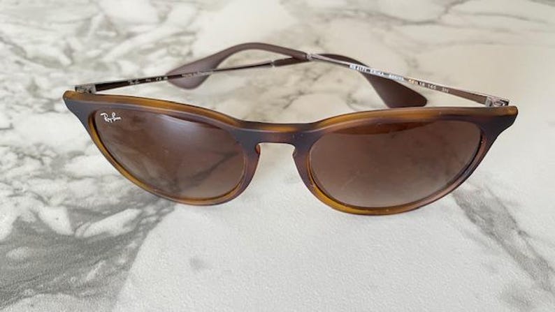 May include: Brown sunglasses with a tortoiseshell frame and gradient lenses. The sunglasses have a classic design with a subtle cat-eye shape. The Ray-Ban logo is visible on the frame.