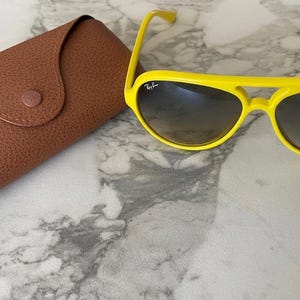 May include: Bright yellow sunglasses with a brown leather case. The sunglasses have a classic aviator shape with a yellow frame and dark lenses. The case is brown with a button closure and the Ray-Ban logo.