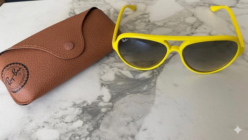 May include: Bright yellow sunglasses with a brown leather case. The sunglasses have a classic aviator shape with a yellow frame and dark lenses. The case is brown with a button closure and the Ray-Ban logo.