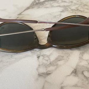 K&ouml;nnte beinhalten: Sonnenbrille mit Schildpattrahmen und dunklen Gl&auml;sern. Die B&uuml;gel sind braun mit silbernen Metallscharnieren. Die Sonnenbrille liegt auf einer Marmoroberfl&auml;che.