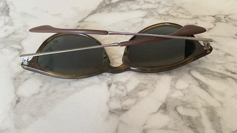 K&ouml;nnte beinhalten: Sonnenbrille mit Schildpattrahmen und dunklen Gl&auml;sern. Die B&uuml;gel sind braun mit silbernen Metallscharnieren. Die Sonnenbrille liegt auf einer Marmoroberfl&auml;che.