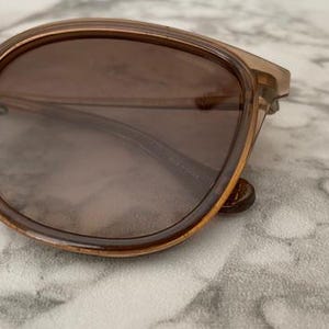 May include: Close-up of sunglasses with brown tinted lenses and a light brown frame. The sunglasses have a rounded shape and a metal bridge. The lenses are dark and provide shade. The sunglasses are resting on a marble surface.