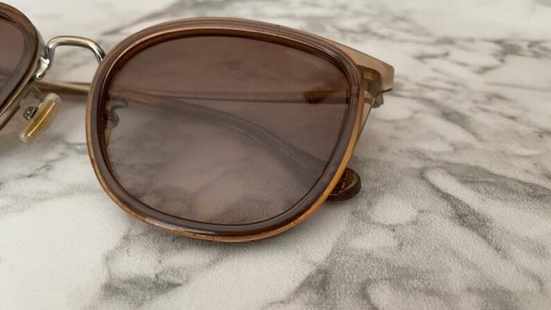 May include: Close-up of sunglasses with brown tinted lenses and a light brown frame. The sunglasses have a rounded shape and a metal bridge. The lenses are dark and provide shade. The sunglasses are resting on a marble surface.