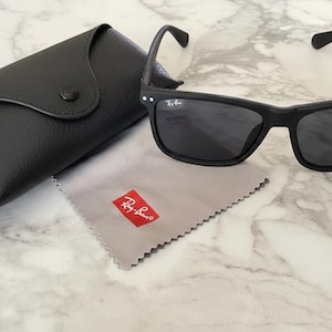 Pu&ograve; includere: Occhiali da sole neri con custodia e panno per la pulizia abbinati. Gli occhiali hanno una montatura rettangolare e lenti scure. La custodia &egrave; nera e il panno per la pulizia &egrave; grigio con un logo rosso. Un accessorio classico.