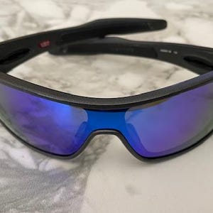 Może przedstawiać: Czarne okulary przeciwsłoneczne z niebieskimi lustrzanymi soczewkami. Okulary mają elegancki, nowoczesny design z czarną oprawką. Soczewki odbijają żywy niebieski kolor. Przeznaczone do użytku na zewnątrz.