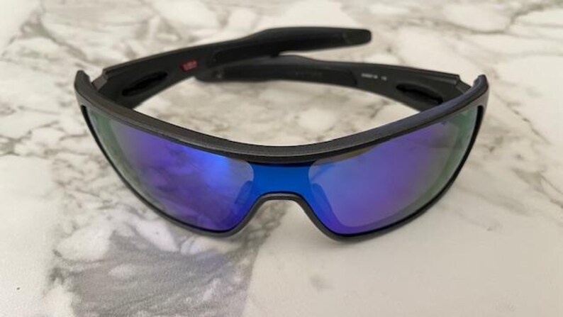 Może przedstawiać: Czarne okulary przeciwsłoneczne z niebieskimi lustrzanymi soczewkami. Okulary mają elegancki, nowoczesny design z czarną oprawką. Soczewki odbijają żywy niebieski kolor. Przeznaczone do użytku na zewnątrz.