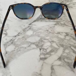 May include: Sunglasses with a tortoiseshell pattern frame and blue-tinted lenses. The sunglasses are resting on a white and gray marble surface. The arms of the sunglasses are dark brown with a tortoiseshell pattern.