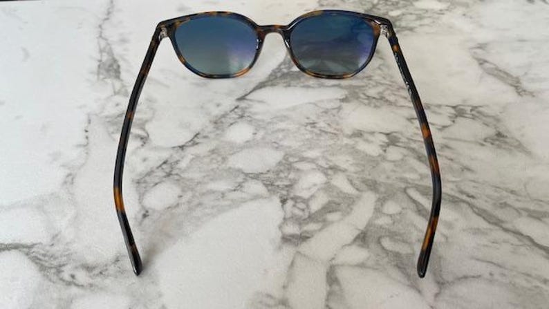 May include: Sunglasses with a tortoiseshell pattern frame and blue-tinted lenses. The sunglasses are resting on a white and gray marble surface. The arms of the sunglasses are dark brown with a tortoiseshell pattern.