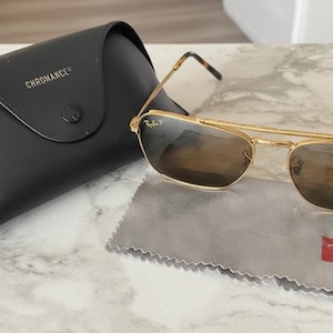Puede incluir: Gafas de sol con montura dorada y lentes marrones, junto a un estuche de cuero negro. El estuche lleva la inscripción "CHROMANCE" y el logotipo de Ray-Ban. También hay un paño de limpieza gris y una etiqueta Ray-Ban.