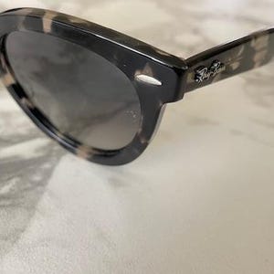 Puede incluir: Gafas de sol carey con lentes oscuras. Las monturas tienen un patr&oacute;n moteado en tonos negro, marr&oacute;n y beige. El logotipo de Ray-Ban es visible en la patilla. Las gafas de sol est&aacute;n sobre un fondo claro.