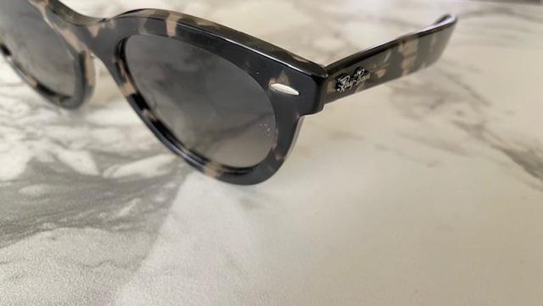 Puede incluir: Gafas de sol carey con lentes oscuras. Las monturas tienen un patr&oacute;n moteado en tonos negro, marr&oacute;n y beige. El logotipo de Ray-Ban es visible en la patilla. Las gafas de sol est&aacute;n sobre un fondo claro.