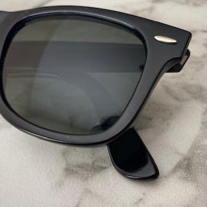 K&ouml;nnte beinhalten: Schwarze Sonnenbrille mit dunklen Gl&auml;sern. Die Sonnenbrille hat ein klassisches, quadratisches Rahmendesign mit gl&auml;nzendem Finish. Ein kleiner silberner Akzent ist an der Fassung in der N&auml;he der Scharniere sichtbar. Die Sonnenbrille liegt auf einer hellen Oberfl&auml;che.