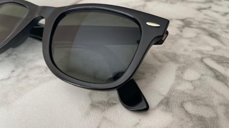 K&ouml;nnte beinhalten: Schwarze Sonnenbrille mit dunklen Gl&auml;sern. Die Sonnenbrille hat ein klassisches, quadratisches Rahmendesign mit gl&auml;nzendem Finish. Ein kleiner silberner Akzent ist an der Fassung in der N&auml;he der Scharniere sichtbar. Die Sonnenbrille liegt auf einer hellen Oberfl&auml;che.