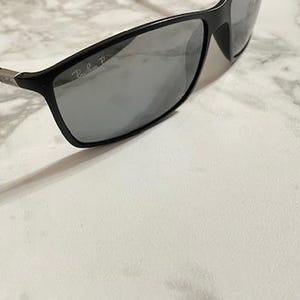 May include: Black rectangular sunglasses with gray lenses. The sunglasses have a sleek, modern design with a matte finish. The Ray-Ban logo is visible on the lens. The frame is black with silver accents on the temples.