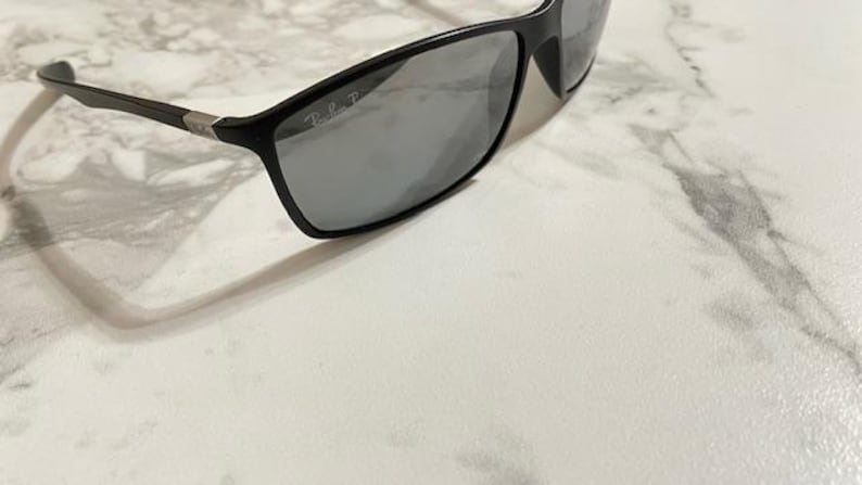 May include: Black rectangular sunglasses with gray lenses. The sunglasses have a sleek, modern design with a matte finish. The Ray-Ban logo is visible on the lens. The frame is black with silver accents on the temples.
