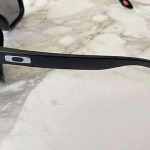May include: Black sunglasses with a rectangular frame and dark lenses. The sunglasses have a sleek, modern design with a silver logo on the temple. The glasses are resting on a white and gray marble surface.