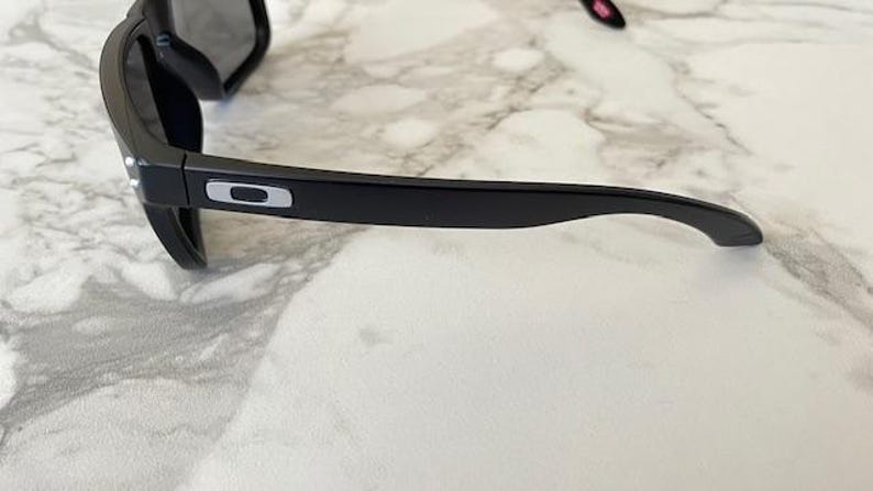 May include: Black sunglasses with a rectangular frame and dark lenses. The sunglasses have a sleek, modern design with a silver logo on the temple. The glasses are resting on a white and gray marble surface.