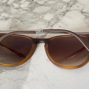 May include: Brown sunglasses with a silver bridge and brown arms. The lenses are a gradient brown color. The sunglasses are resting on a white and gray marble surface.