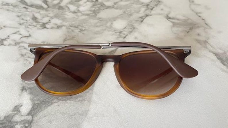 May include: Brown sunglasses with a silver bridge and brown arms. The lenses are a gradient brown color. The sunglasses are resting on a white and gray marble surface.