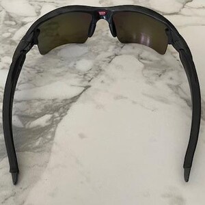Puede incluir: Gafas de sol negras con montura curvada y lentes tintadas. Las gafas de sol tienen un peque&ntilde;o detalle rojo y blanco en el puente. Las patillas de las gafas son curvas y se extienden hacia afuera.