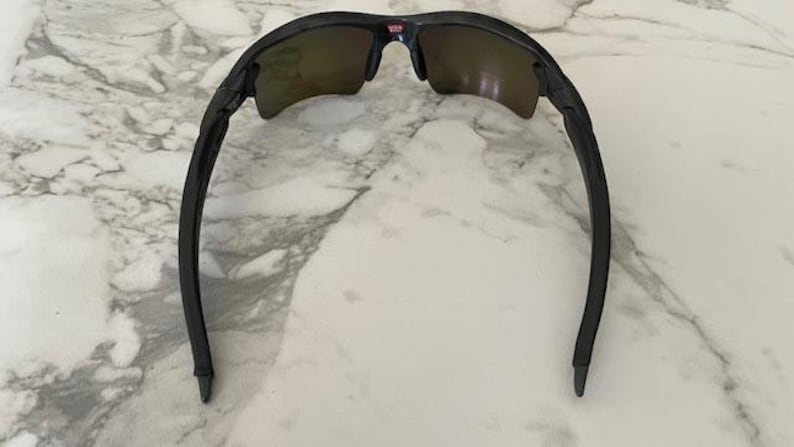 Puede incluir: Gafas de sol negras con montura curvada y lentes tintadas. Las gafas de sol tienen un peque&ntilde;o detalle rojo y blanco en el puente. Las patillas de las gafas son curvas y se extienden hacia afuera.