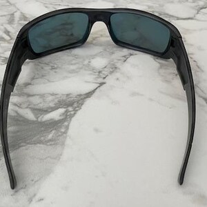 Puede incluir: Gafas de sol negras con lentes de color azul. Las gafas de sol tienen un dise&ntilde;o curvo y deportivo con monturas y patillas gruesas. Las lentes son rectangulares y las monturas son de color negro mate. Sobre fondo de m&aacute;rmol blanco.