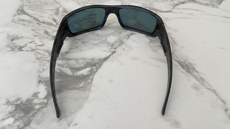 Puede incluir: Gafas de sol negras con lentes de color azul. Las gafas de sol tienen un dise&ntilde;o curvo y deportivo con monturas y patillas gruesas. Las lentes son rectangulares y las monturas son de color negro mate. Sobre fondo de m&aacute;rmol blanco.
