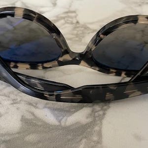 Puede incluir: Gafas de sol con montura con estampado de carey y lentes azules. Las gafas de sol est&aacute;n plegadas y descansan sobre una superficie de m&aacute;rmol. Las patillas de las gafas de sol tienen el nombre de la marca impreso.
