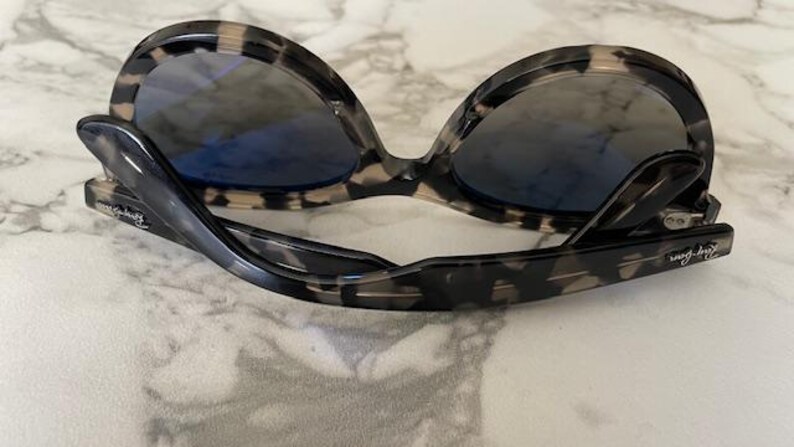 Puede incluir: Gafas de sol con montura con estampado de carey y lentes azules. Las gafas de sol est&aacute;n plegadas y descansan sobre una superficie de m&aacute;rmol. Las patillas de las gafas de sol tienen el nombre de la marca impreso.