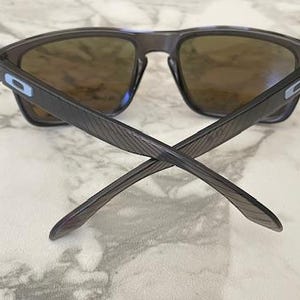May include: Dark gray sunglasses with brown lenses. The arms of the sunglasses are crossed, showing the textured design. The frames have a rectangular shape. The lenses are a brown tint, and the sunglasses have a modern, sporty look.