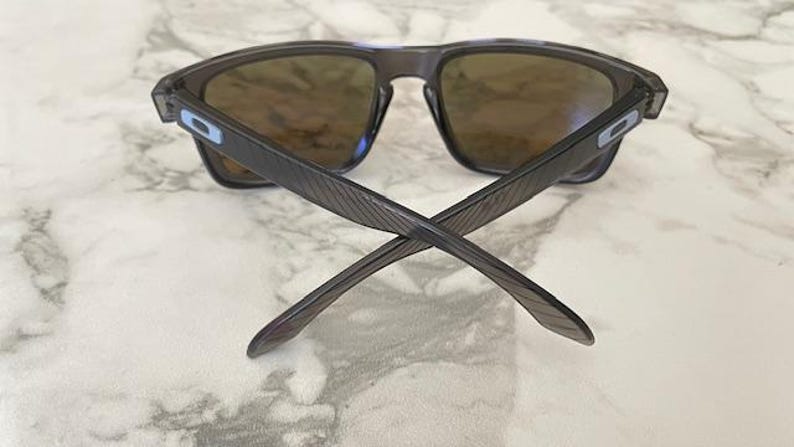 May include: Dark gray sunglasses with brown lenses. The arms of the sunglasses are crossed, showing the textured design. The frames have a rectangular shape. The lenses are a brown tint, and the sunglasses have a modern, sporty look.