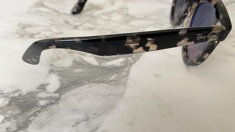 Puede incluir: Gafas de sol carey negras y marrones con lentes azules. El logotipo de Ray-Ban es visible en la patilla. Colocadas sobre una superficie de m&aacute;rmol blanco y gris.